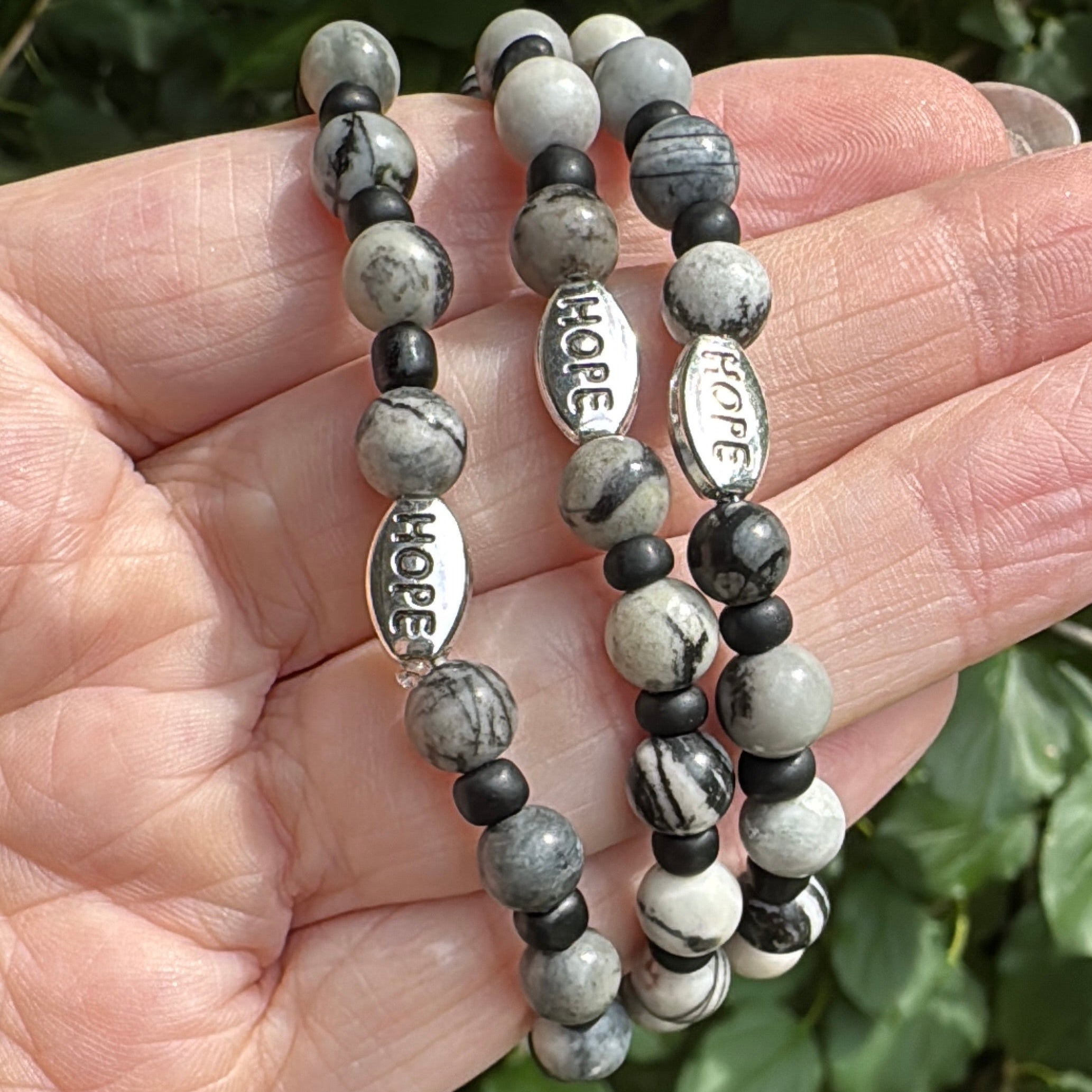 Three beaded stretch bracelets with gray and white marbled beads, small black beads, and silver nameplates spelling HOPE, LOVE, and BRITISH COLUMBIA, held in a hand against a background of green leaves.