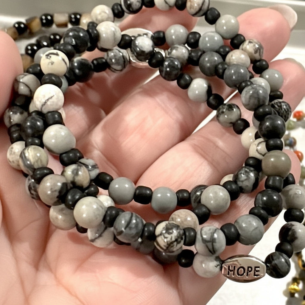 A close-up of a hand holding a coiled beaded bracelet made of black, white, and gray marbled beads, with a small round charm engraved with the word 'HOPE'.