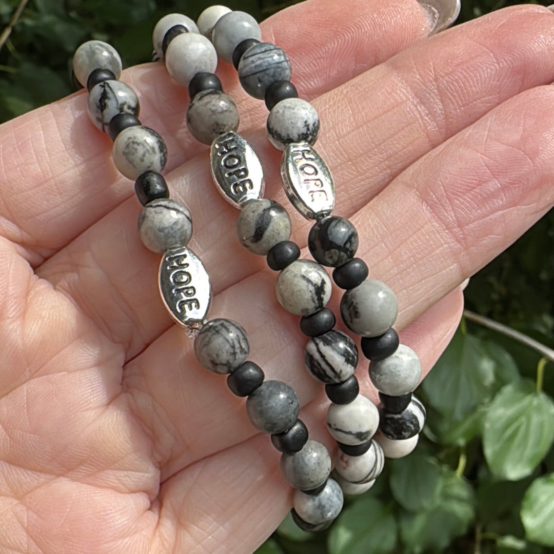 Three beaded stretch bracelets with gray and black marbled beads and silver 'HOPE' charms, held in a hand against a background of green leaves.