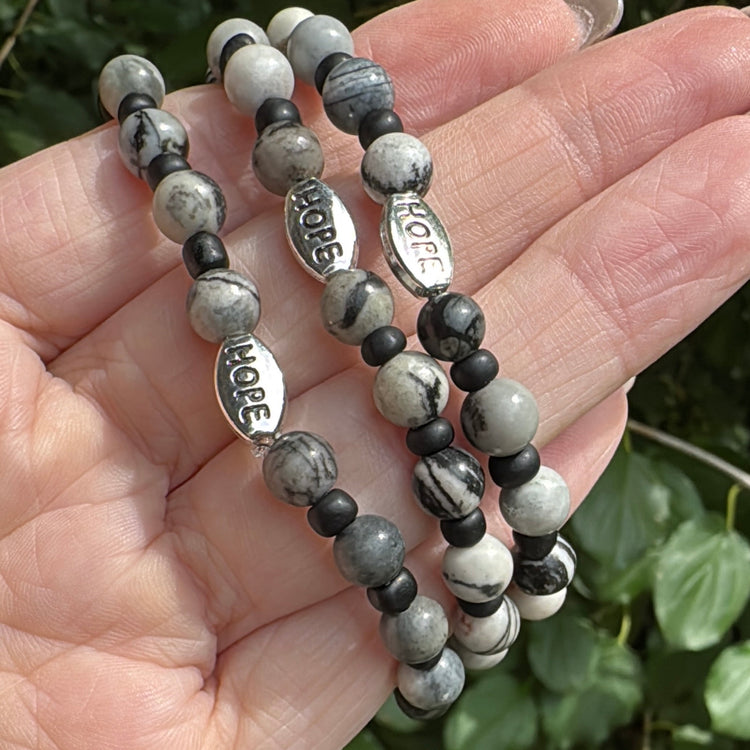 Three beaded stretch bracelets with gray and black marbled beads and silver 'HOPE' charms, held in a hand against a background of green leaves.