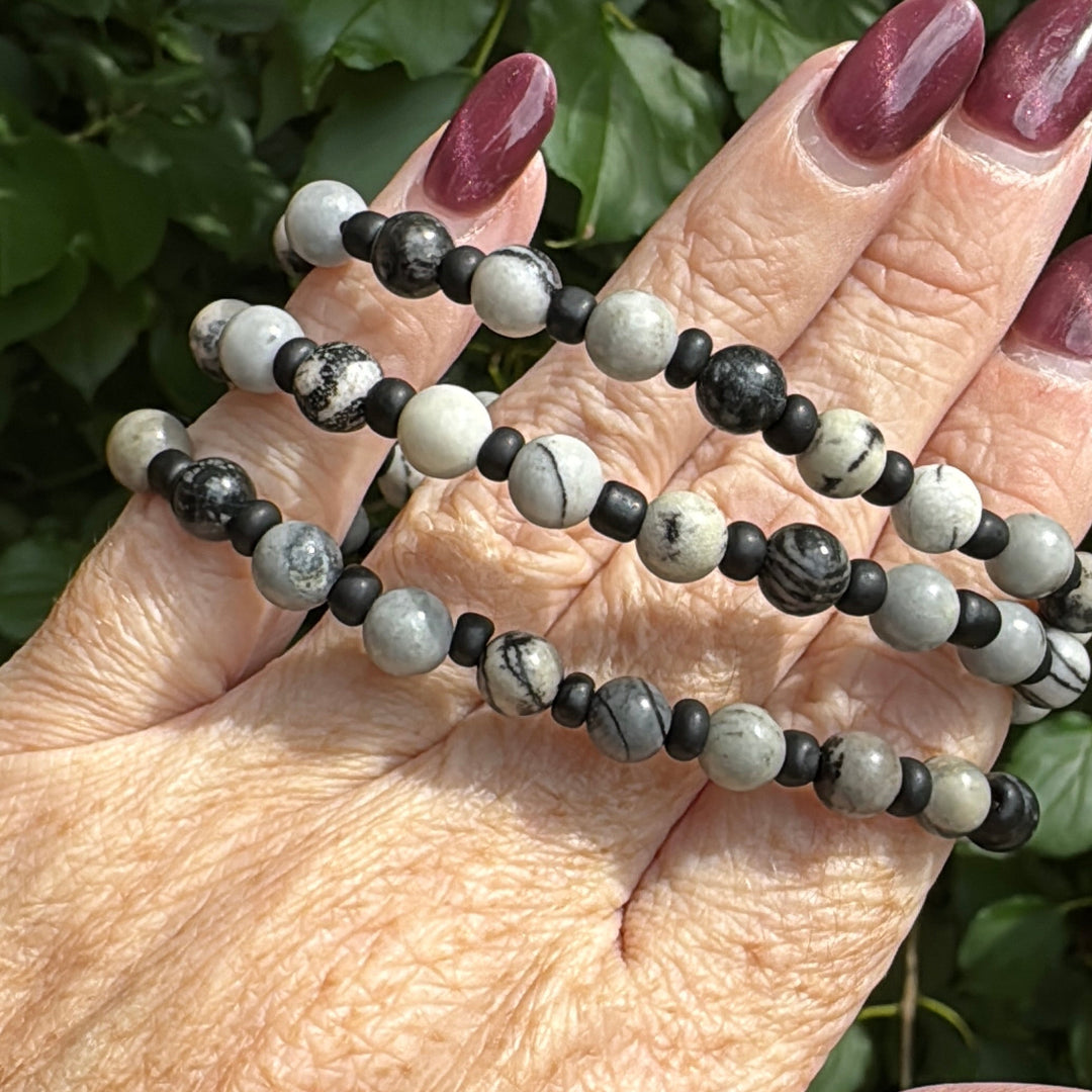 A close-up of a person's hand holding three beaded strands, likely prayer beads or a necklace, featuring black and white marbled beads, with green foliage in the background.