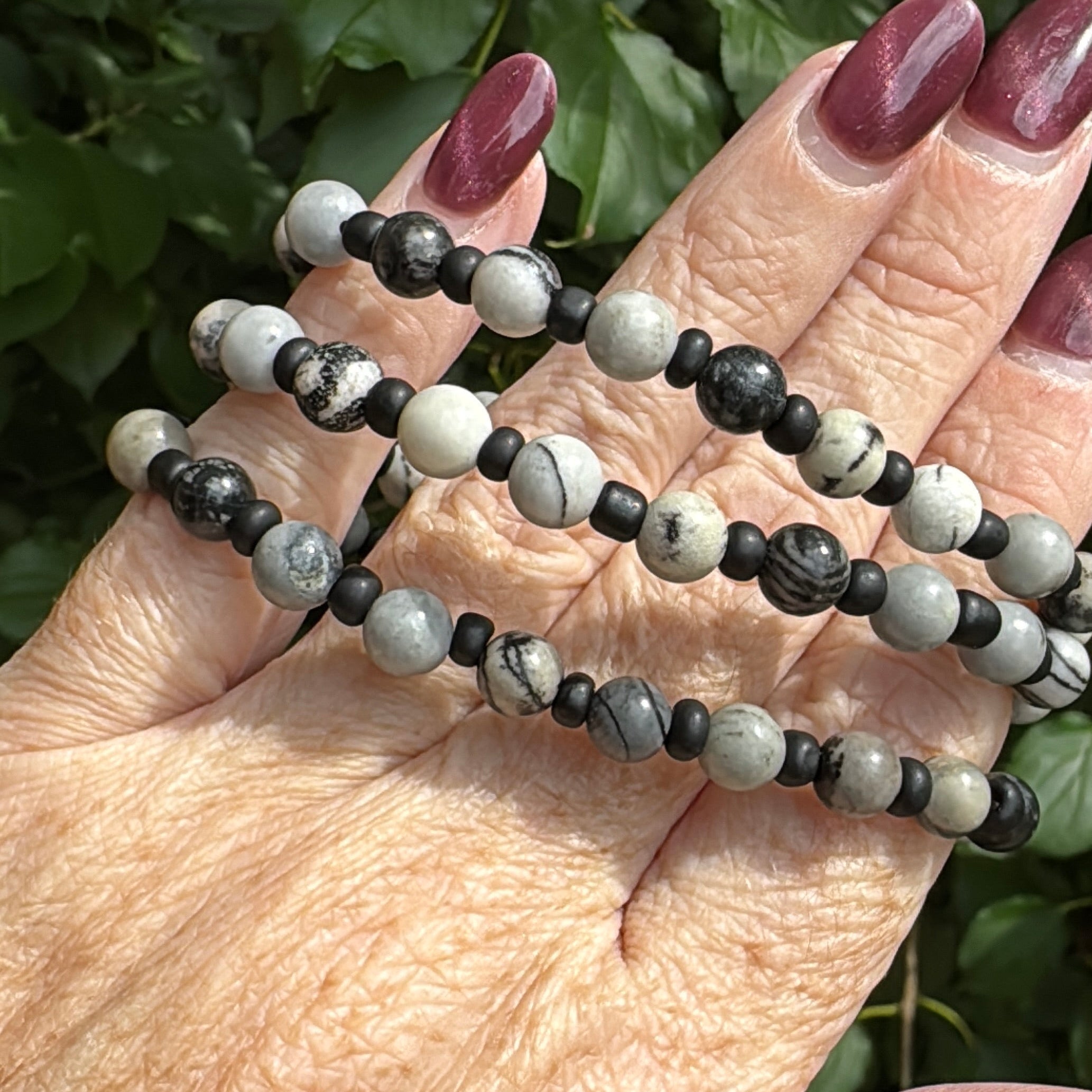 A close-up of a person's hand holding three beaded strands, likely prayer beads or a necklace, featuring black and white marbled beads, with green foliage in the background.
