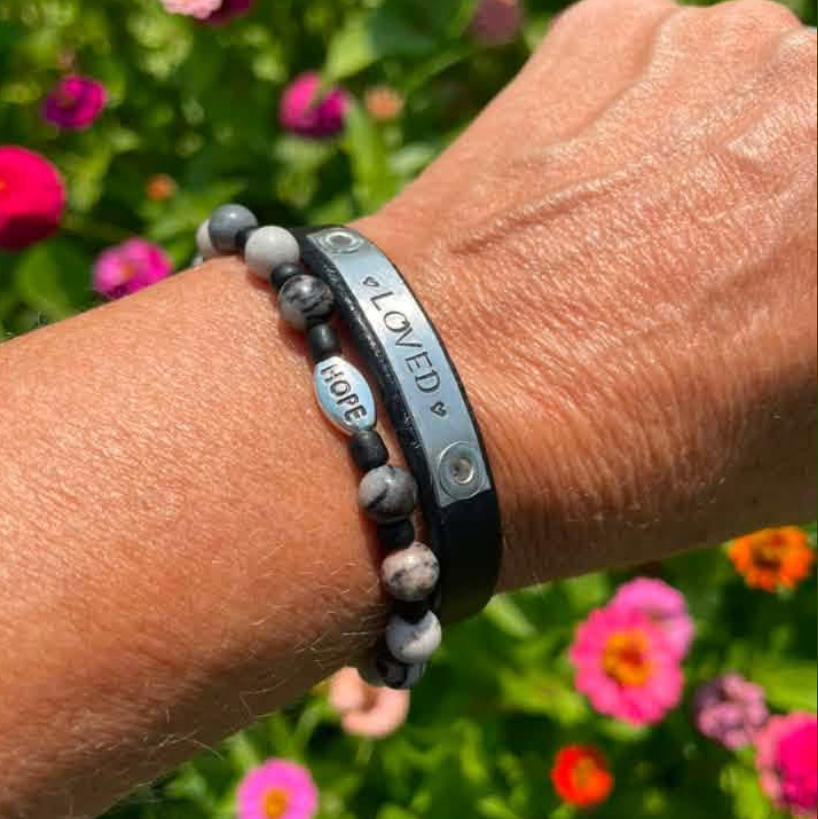 A close-up of a person's wrist wearing three beaded and metal bracelets outdoors, with a blurred background of green foliage and pink flowers. The bracelets include a black band engraved with 'LOVED' and 'HOPE', a silver-toned engraved band, and a gray and black beaded bracelet.